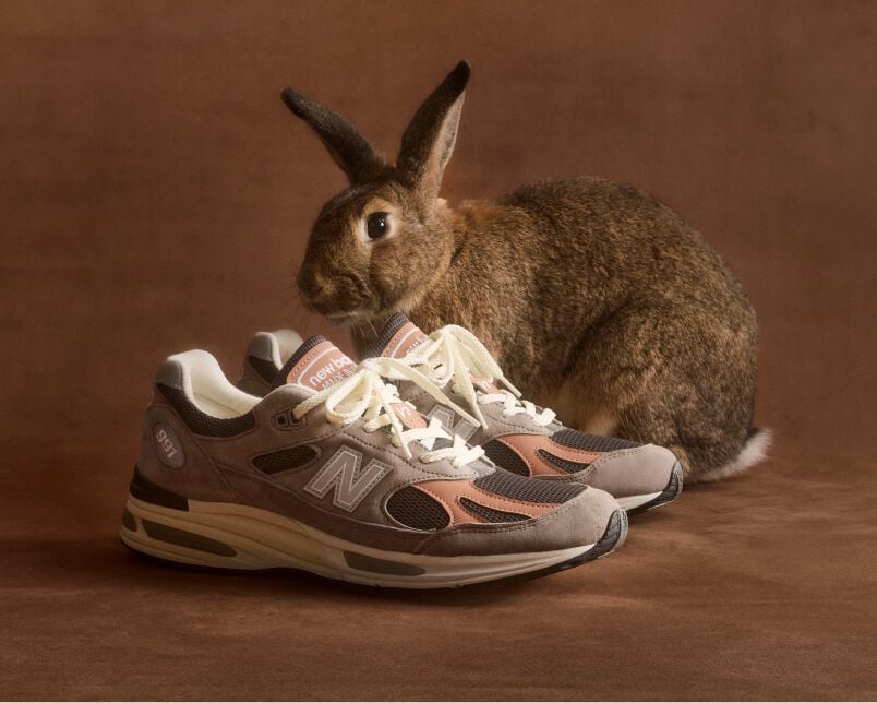 A pair of New Balance Made in UK sneakers in front of a rabbit in a brown textured studio environment. 