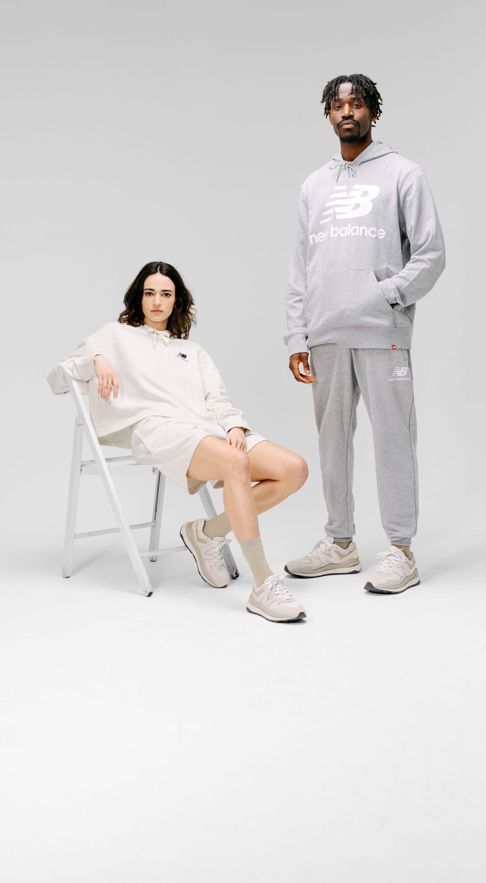 Man and woman wearing grey New Balance apparel and footwear in a grey studio environment.