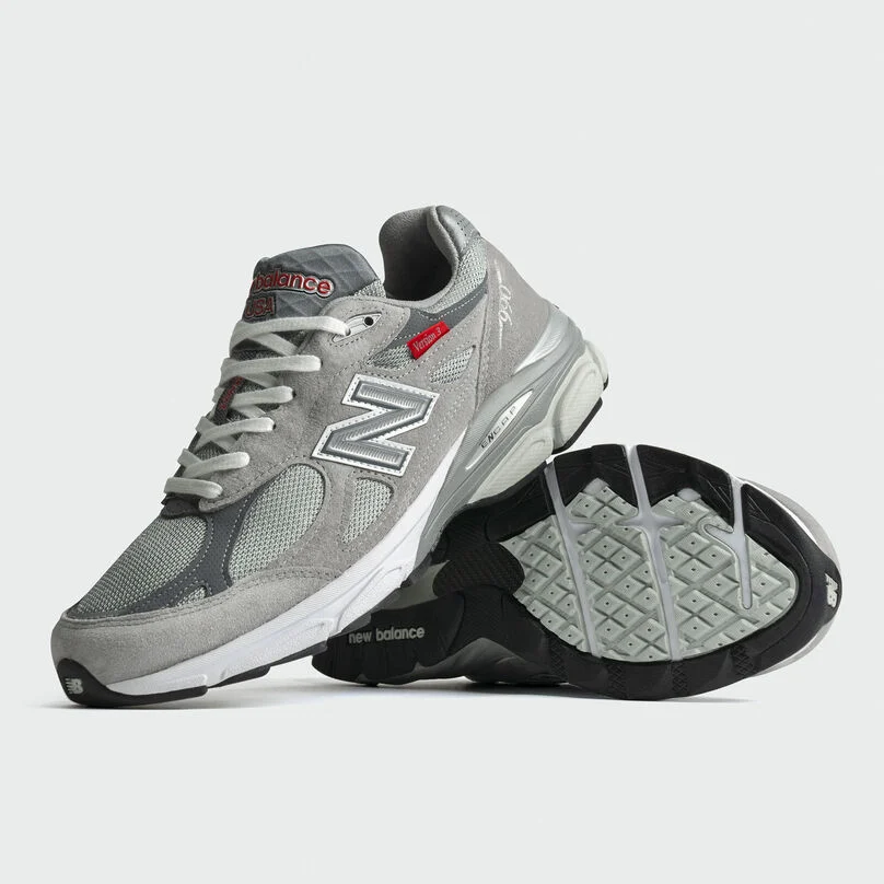 Pair of grey 990v3 sneakers stacked on each other in a grey studio environment.