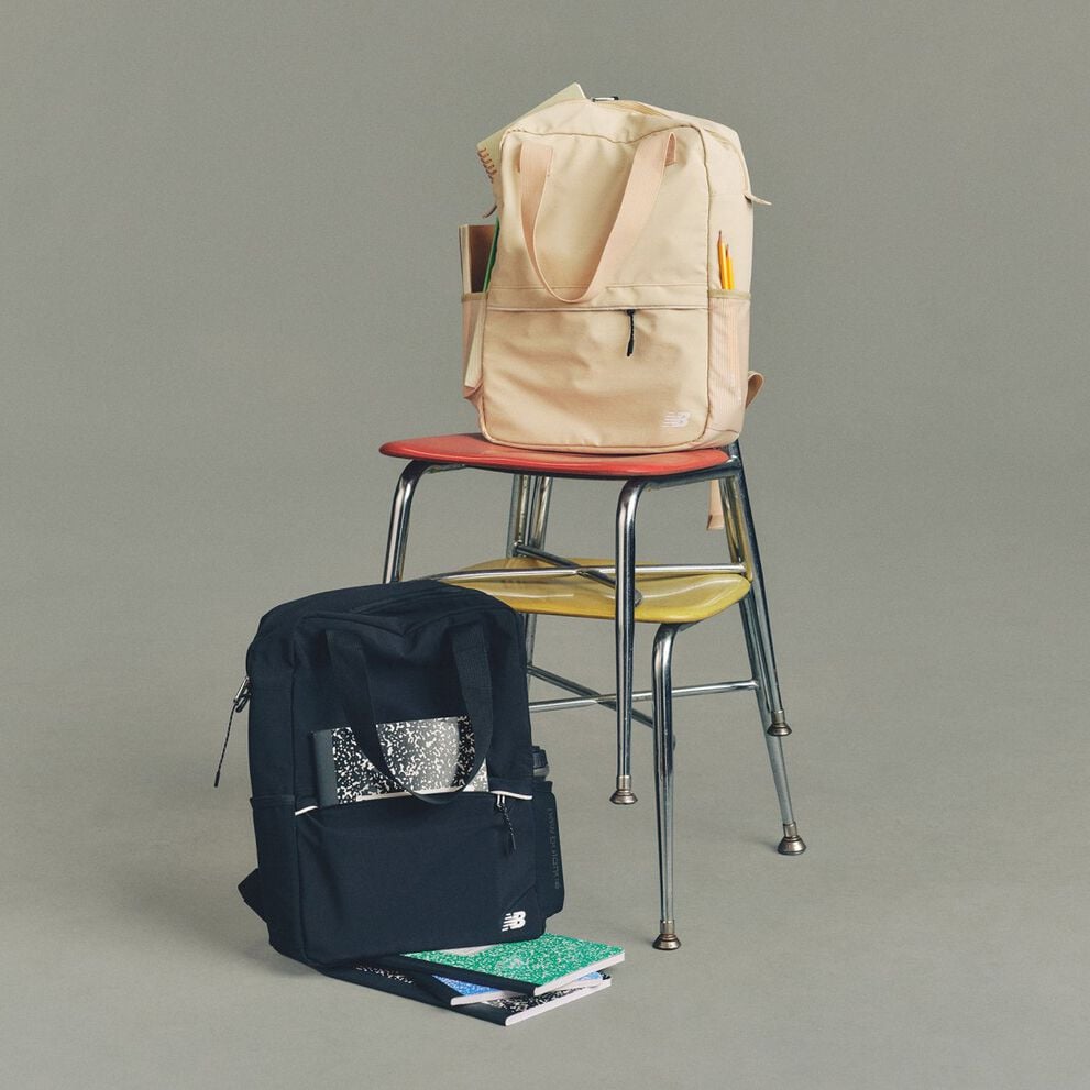 Studio image of a pair of cream-colored New Balance sneakers placed on a dark wooden folding chair, with a second matching chair folded and leaned behind it.