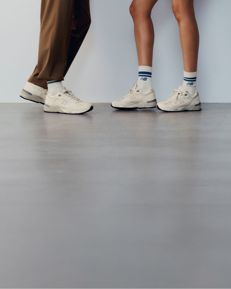 Knee down view of a man and woman wearing New Balance Made in UK sneakers standing on a concrete floor against a grey wall. 