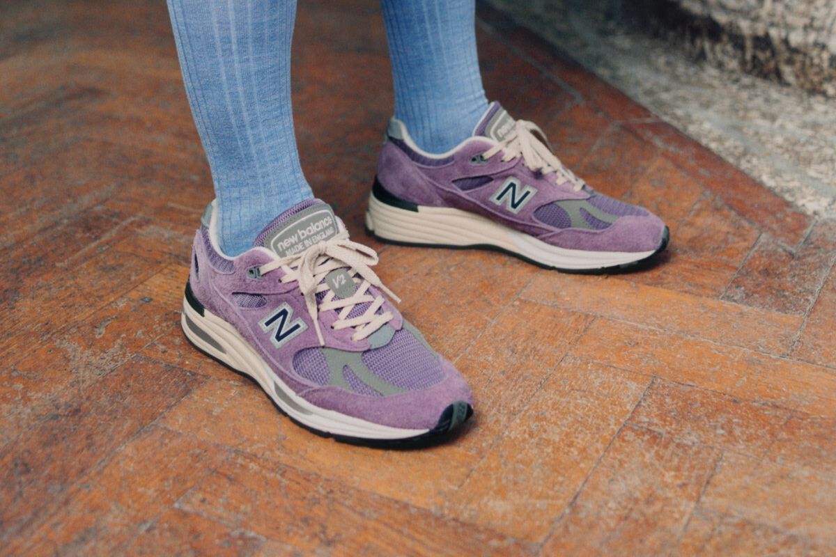 A pair of New Balance Made in UK sneakers standing on a wooden floor with textured green walls and a stone pillar in the background.