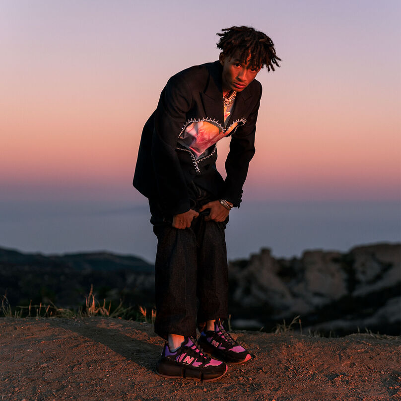 NB ambassador Jaden Smith bending forward with his pant legs raised above his ankles. He is wearing a black blazer embroidered with an orange and pink heart with black pants and black NB Vision Racer sneakers that have pink and orange accents. He is framed by a scenic sunset overlooking the ocean.